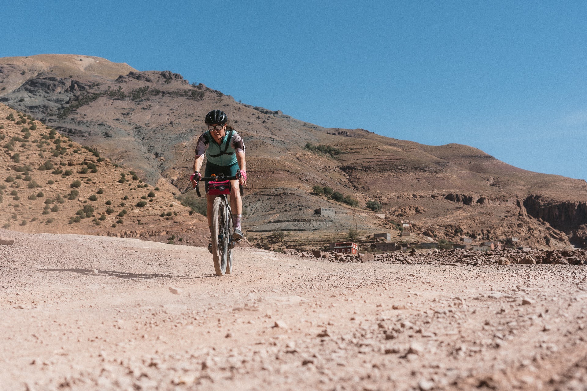 Cyclist riding a bike on a rocky dirt road with arid mountains in the background under a clear blue sky.