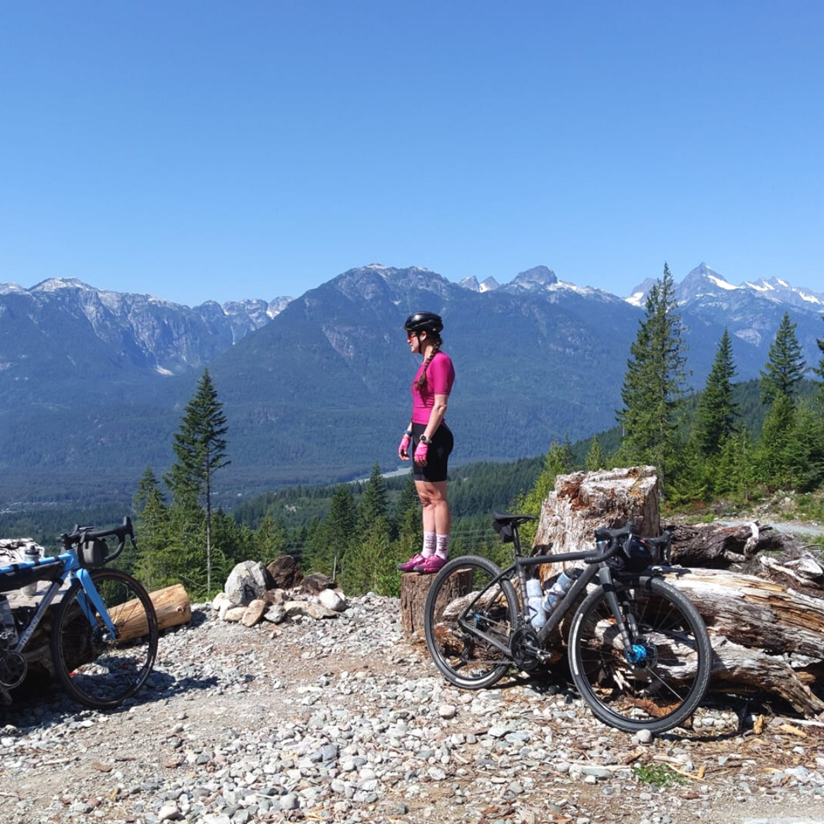 Cyclist in pink outfit standing on a tree stump on a rocky trail with mountain bikes and a scenic mountain range in the background under a clear blue sky.