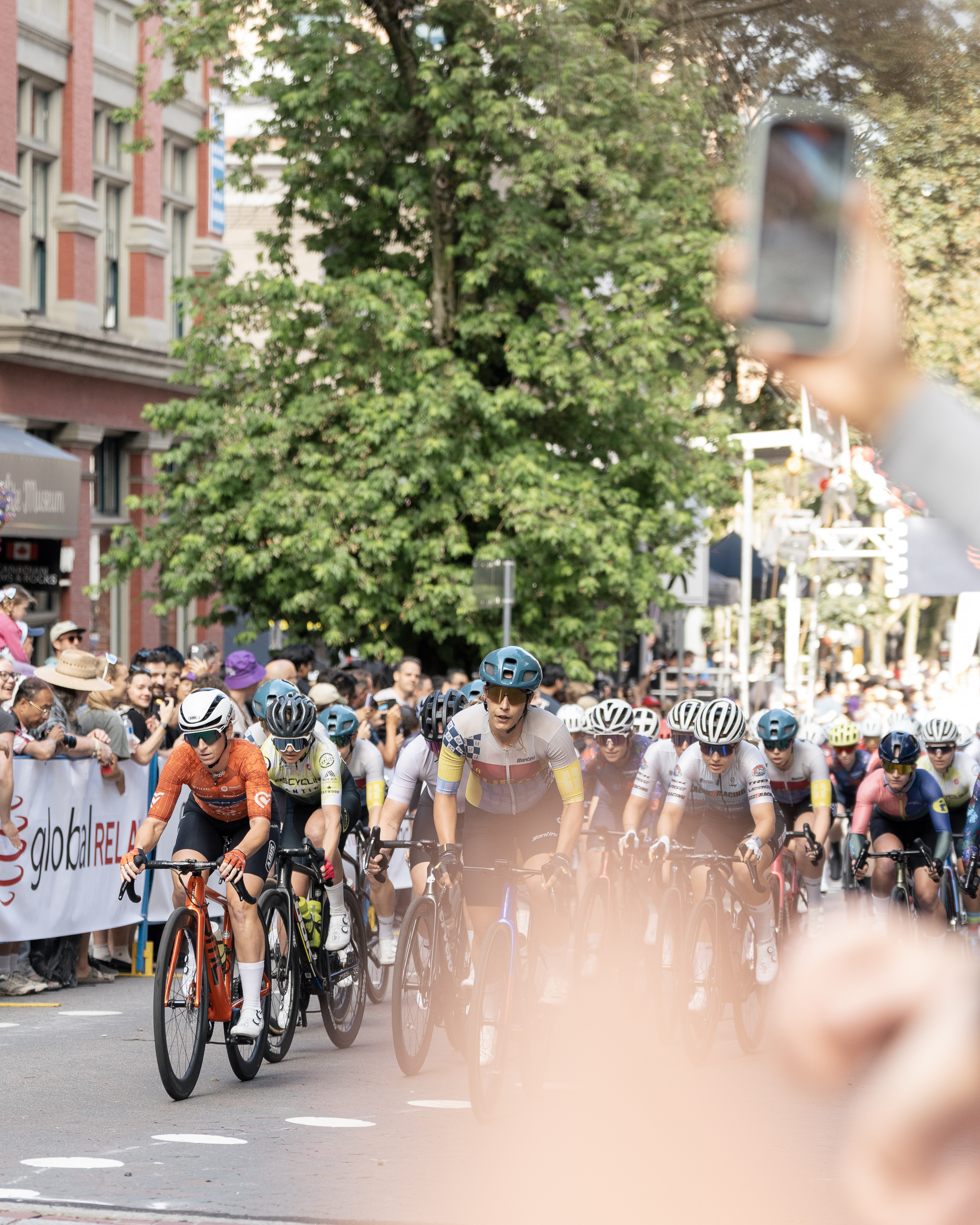 Group of cyclists racing on a city street with spectators and a person holding a phone in the foreground.