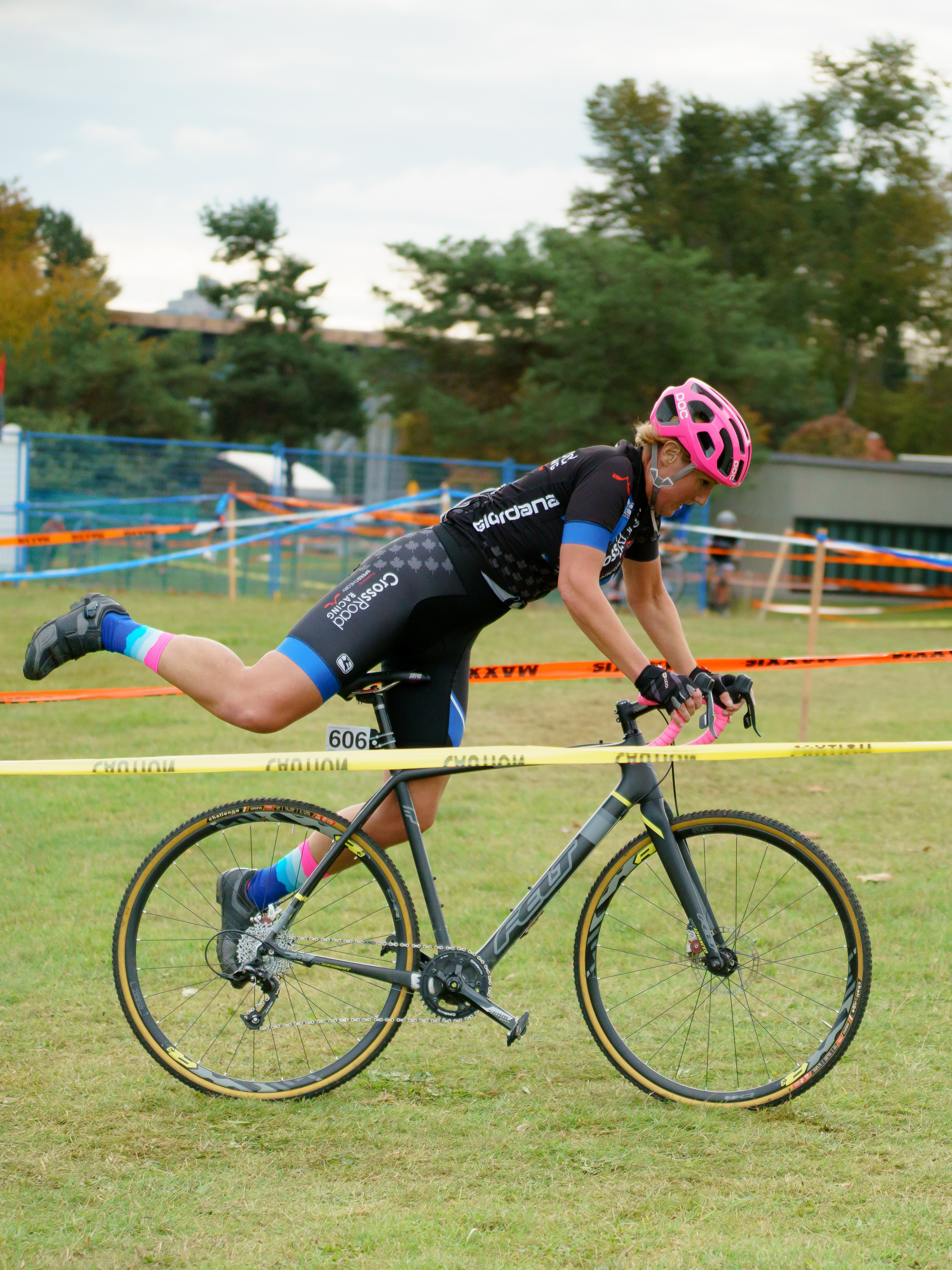 Cyclist in black and blue gear with pink helmet and socks mounting a gray bike over grassy terrain marked with caution tape.