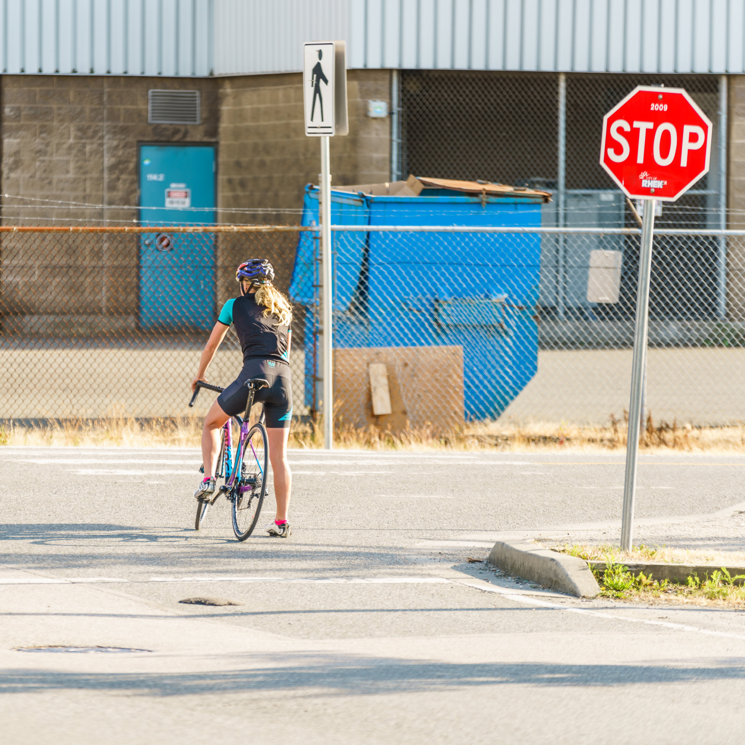 Cyclist wearing a helmet and black cycling outfit riding a bicycle on a street near a stop sign and pedestrian crossing sign with a blue dumpster and chain-link fence in the background.